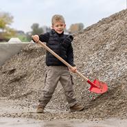 Ein lächelnder Junge in Outdoor-Kleidung hält die Frankfurter Schaufel "Mini" für Kinder, mit Stiel, 107cm, vor einem großen Kieshaufen stehend.