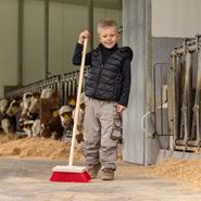 Ein lächelnder Junge hält den Straßenbesen "Mini" für Kinder mit Stiel (110 cm) in einer Scheune, in der im Hintergrund Kühe zu sehen sind.