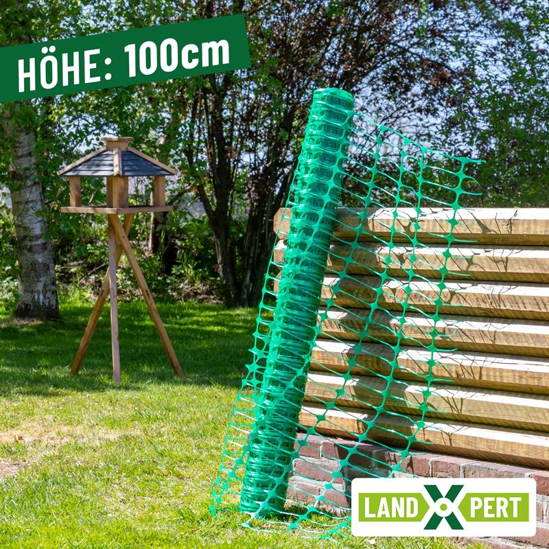 Großansicht der Bildergalerie Der LandXpert 50x1m Basic Begrenzungszaun, ein flexibler grüner Kunststoff-Gartenzaun (100 cm hoch), lehnt im Freien an einem Holzzaun, im Hintergrund ist ein Vogelfutterhaus zu sehen.