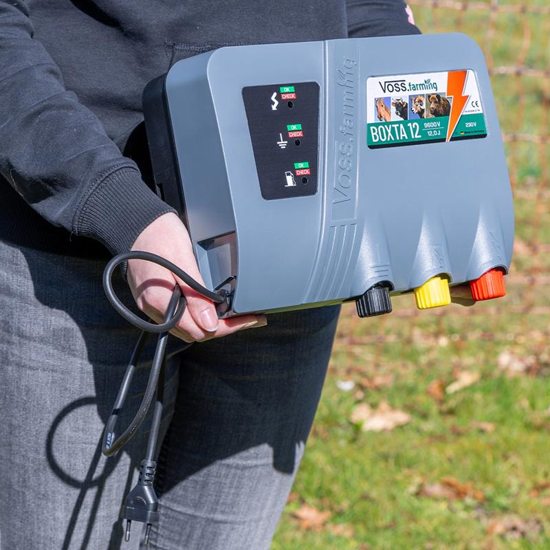 Eine Person hält das VOSS.farming Weidezaungerät „BOXTA 12“ 230V im Außenbereich und präsentiert dessen Kabel und farbige Anschlüsse.