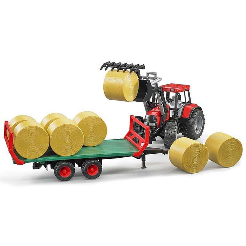 Ein BRUDER Ballentransportanhänger (1:16 Spielzeug-Anhänger, ab 3 Jahren, inkl. 8 Rundballen) lädt mit rotem Traktor Heuballen auf einen grünen Anhänger, während weitere Ballen in der Nähe liegen.