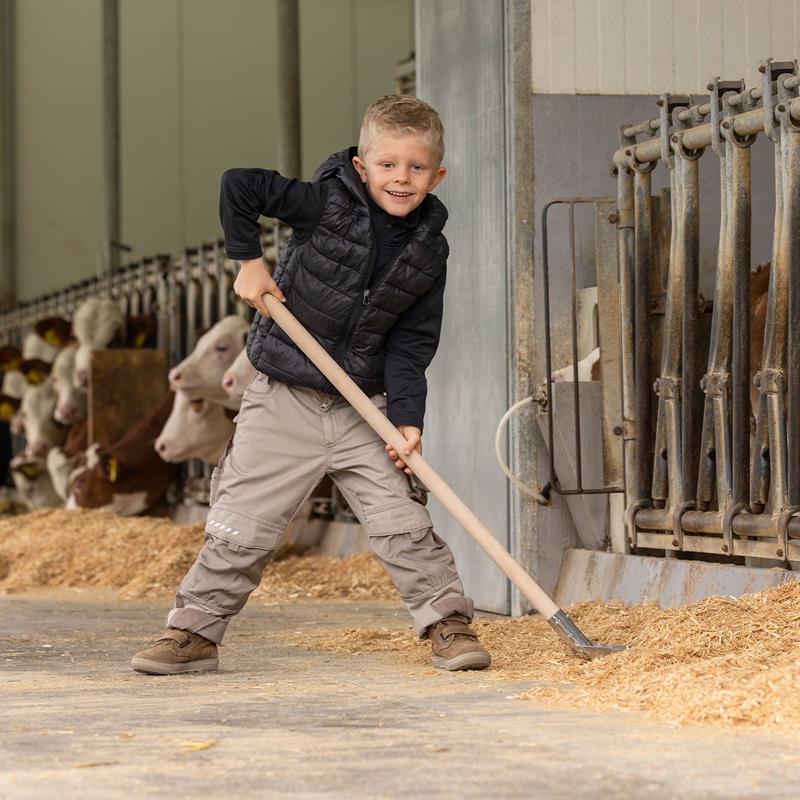 Großansicht der Bildergalerie Ein lächelnder Junge steht in einer Scheune und benutzt den Aluschaufel "Mini" für Kinder, mit Stiel, 107cm, während hinter ihm Kühe fressen.