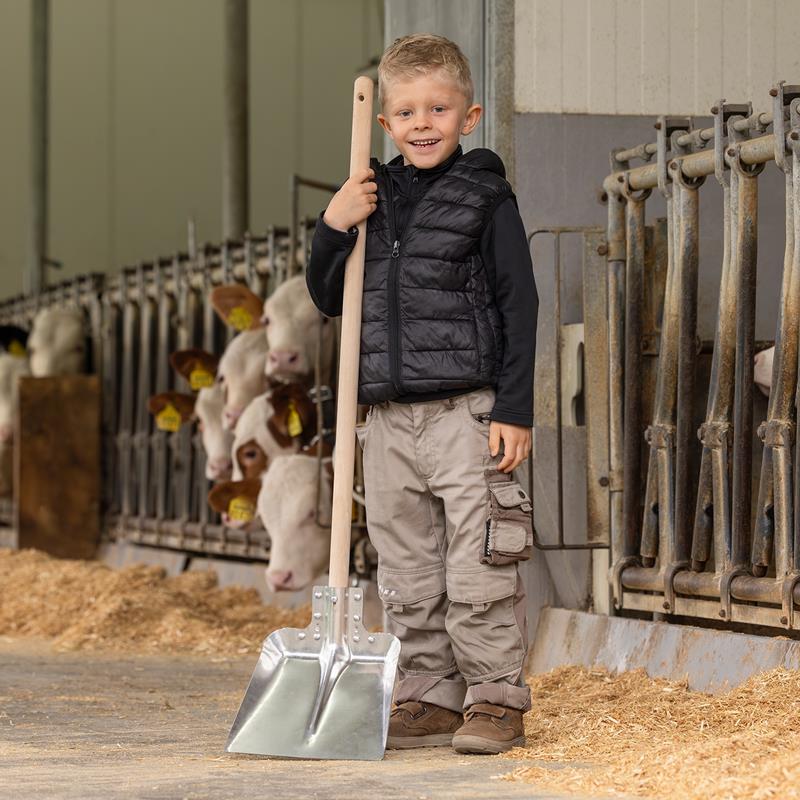 Großansicht der Bildergalerie Ein lächelnder Junge hält den Aluschaufel "Mini" für Kinder mit einem 107 cm langen Stiel in einer Scheune, während im Hintergrund Kühe fressen.
