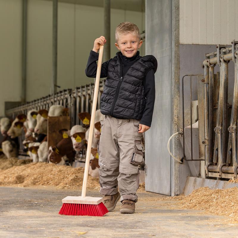 Großansicht der Bildergalerie Ein lächelnder Junge hält den Straßenbesen "Mini" für Kinder mit Stiel (110 cm) in einer Scheune, in der im Hintergrund Kühe zu sehen sind.